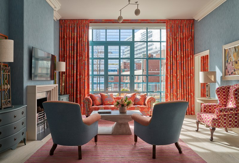 A living area. Two blue and orange armchairs in front of a coffee table opposite an orange 'Chainstitch' sofa which matches curtains on the windows behind. On the left, a fireplace, TV and two chest of drawers either side with lamps on. On the right, pink and white checkered armchair and a side table with a lamp on.