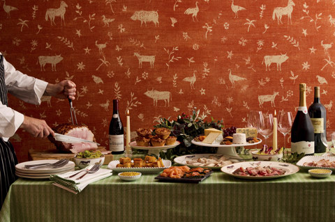 This image shows a festive grazing table featuring a selection of seasonal dishes, to promote Brumus festive offering at Haymarket Hotel. The Table is covered in a green Shop Kit Kemp table cloth and includes a carved glazed ham, smoked salmon, cold meats, a cheese platter, mince pies and accompaniments, styled with foliage, candles and wine against a warm patterned wallpaper by Kit Kemp at Haymarket Hotel.