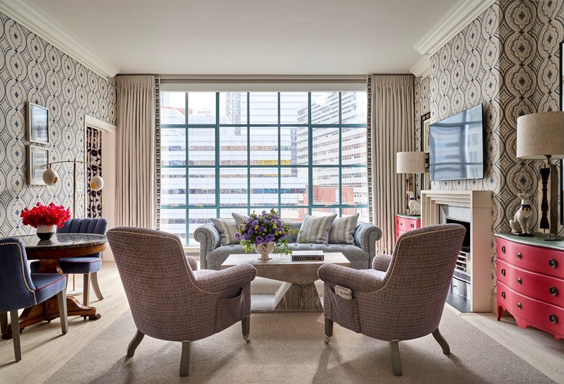Two blue and red patterned armchairs in front of a coffee table opposite a blue sofa. On the right, two dark blue chairs, a table and a standing lamp. On the left, a fireplace and TV on the wall, a red chest of drawers and dog lamp on either side.