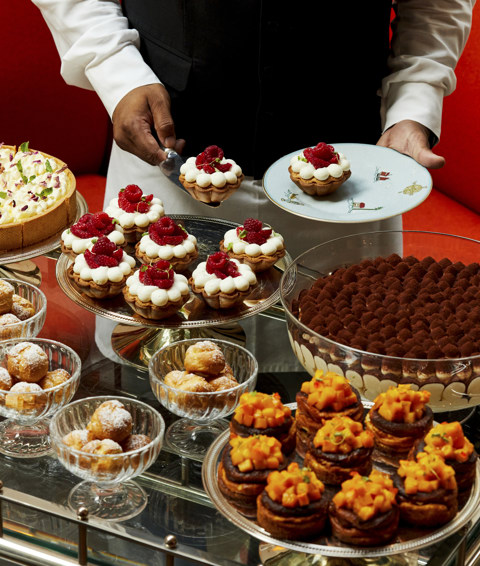 A portrait image taken of The Long Lunch at Ham Yard Bar & Restaurant. The image shows a luxurious dessert trolley laden with an assortment of intricately presented pastries and cakes. A server in formal attire is plating a raspberry tart.