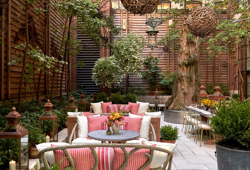 Wide shot from the left of the curtain wall of the bar, of the lounge sofas and chairs  in the sculpture garden at the Crosby Street Hotel and a large stone table on the right of the shot behind some plant pots and in front of a large tree.