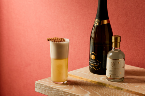 An image of a yellow cocktail in a tall glass, frothy on top.  Next to a bottle of Spirit of Honey, and Northumberland Wildflower sparkling mead 