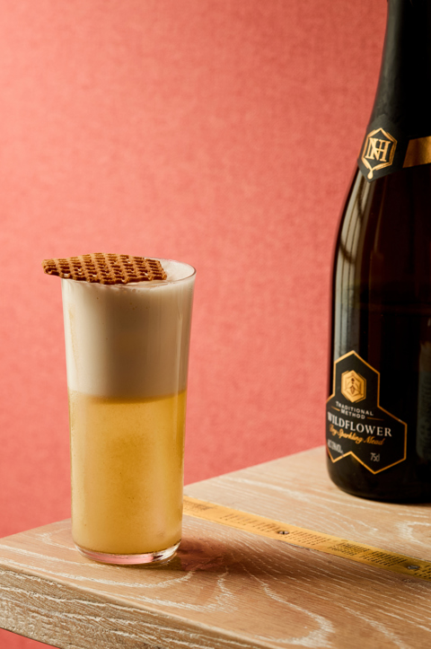 An image of a yellow cocktail in a tall glass, frothy on top. Next to a bottle of Spirit of Honey, and Northumberland Wildflower sparkling mead
