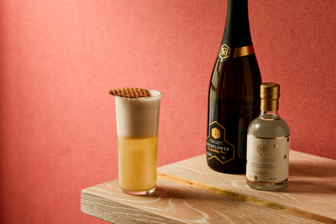 An image of a yellow cocktail in a tall glass, frothy on top. Next to a bottle of Spirit of Honey, and Northumberland Wildflower sparkling mead