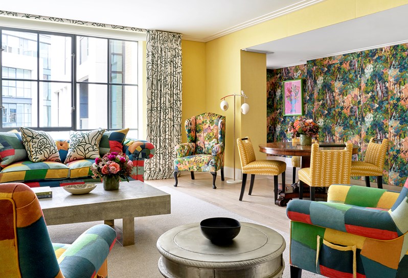 A living area of a suite. The room has yellow walls. A sofa and two armchairs are upholstered in a multi-coloured 'Zig Zag' pattern. On the right, there is a dining table with yellow chairs, in front of a multi-coloured mural.