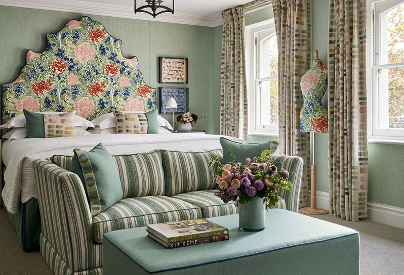 Diagonal bedroom shot a junior suite with plain light blue walls and colourful nature drawings on the headboard and mannequin. There is also  a sofa and a long chair in similar blue, beige and green tones.