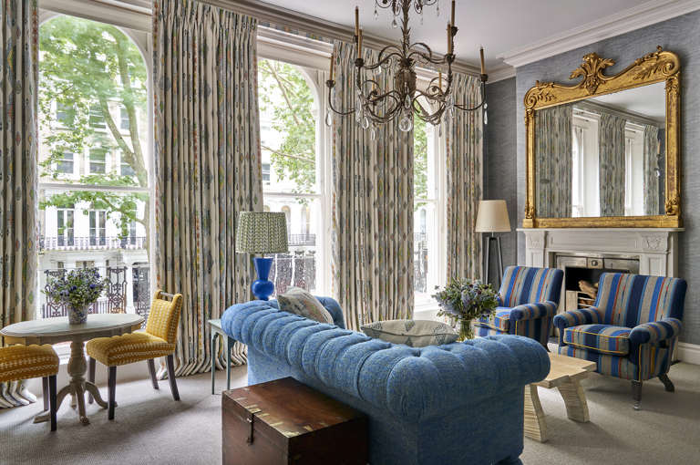 Seating area and fireplace shot of room 101, the Knightsbridge Suite at Knightsbridge Hotel. The image shows a diagonal shot from the bed area across the seating area, showing the blue sofa in the middle of the room facing towards two blue striped armchairs with a low coffee table in the center. A fireplace sits behind the armchair. 