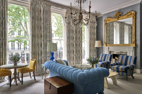 Seating area and fireplace shot of room 101, the Knightsbridge Suite at Knightsbridge Hotel. The image shows a diagonal shot from the bed area across the seating area, showing the blue sofa in the middle of the room facing towards two blue striped armchairs with a low coffee table in the center. A fireplace sits behind the armchair.