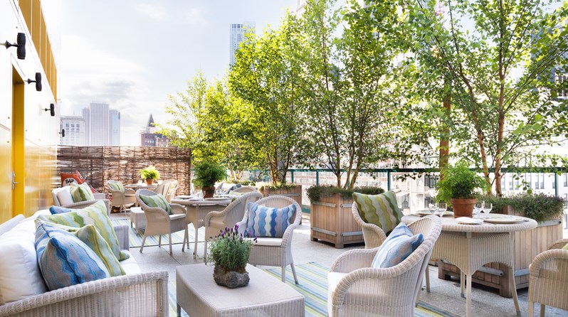 A roof terrace filled with white wicker outdoor sofas, coffee tables, dining tables and chairs. The sofas and chairs display green and blue patterned outdoor cushions. On the tables, there are small potted plants and dining arrangements. The edge of the balcony is lined with small trees.